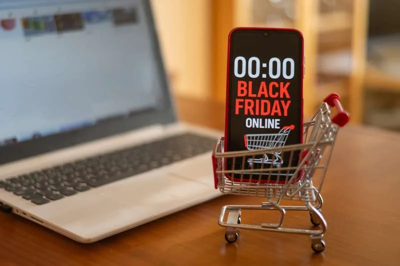 A phone showing a Black Friday countdown, in a small shopping cart beside a laptop